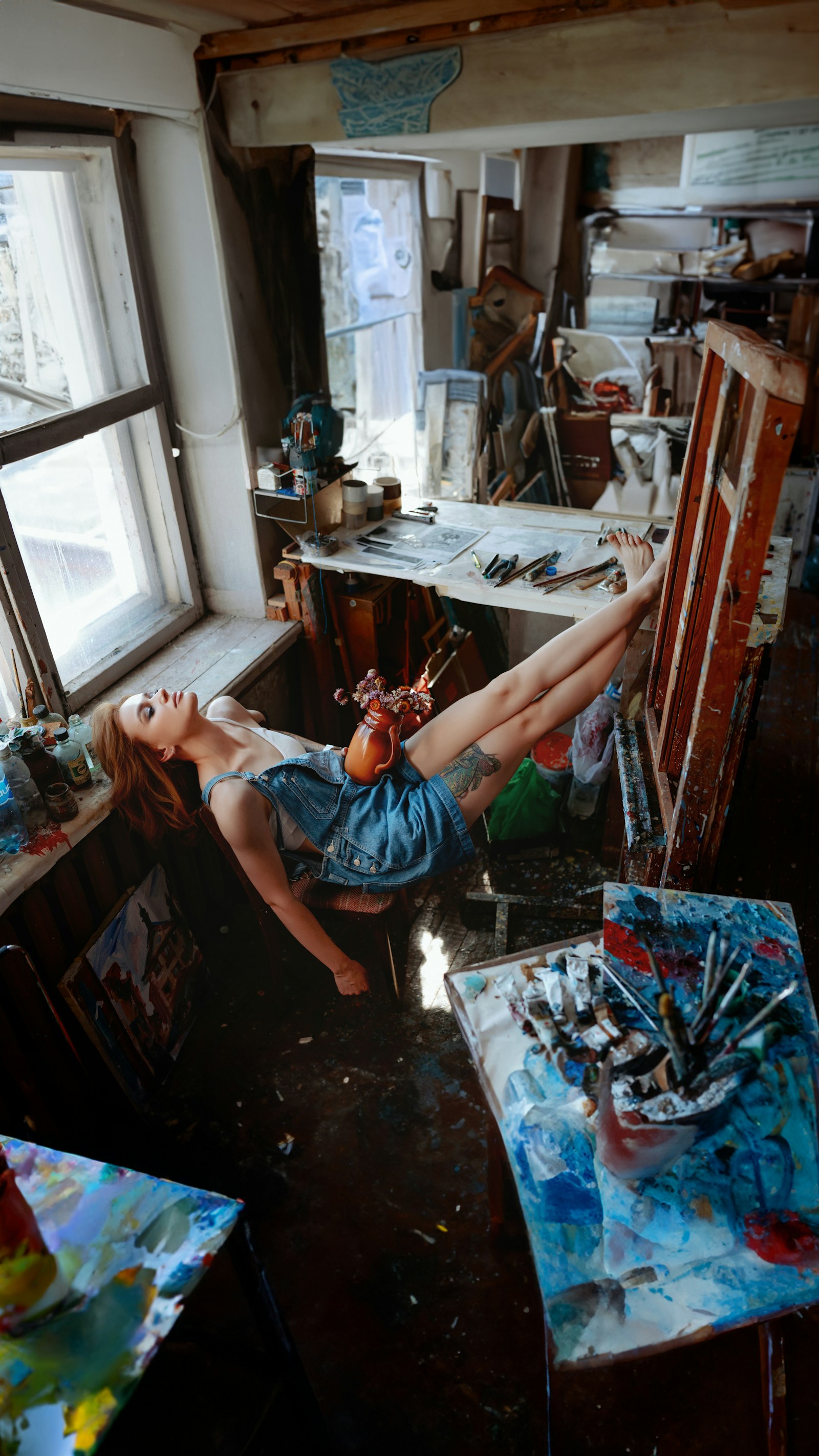 A woman reclining in an art studio, feet resting against artwork and a vase balanced on her lap